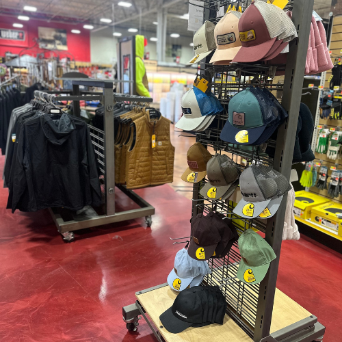 Carhartt-branded hats displayed in the foreground, with Carhartt brown and black vests and a black jacket hanging on a rack behind them inside Peter's True Value Hardware.